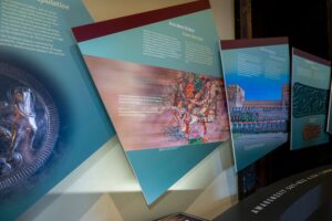 Mehrere schrÃ¤g angebrachte Ausstellungstafeln mit deutsch- und englischsprachigen Infotexten, Illustrationen und historischen Abbildungen, darunter eine MetallprÃ¤gung, eine Zeichnung von Reitern und ein Foto eines GebÃ¤udes. (Text KI-unterstÃ¼tzt generiert)