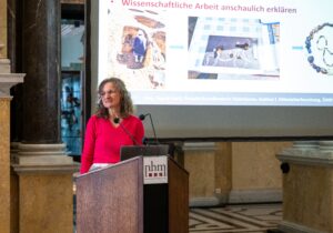 Frau in pinkem Oberteil spricht an einem Rednerpult mit Mikrofonen vor einer groÃen ProjektionsflÃ¤che in einem Saal mit SÃ¤ulen. Auf der Leinwand sind Bilder und der Text âWissenschaftliche Arbeit anschaulich erklÃ¤renâ zu sehen. (Text KI-unterstÃ¼tzt generiert)
