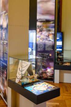 Museumsraum mit Ausstellungsvitrine, Wandtafeln und Infotexten zu ErnÃ¤hrung, Kultur und Genetik sowie mehreren archÃ¤ologischen Objekten in Glasvitrinen. (Text KI-unterstÃ¼tzt generiert)