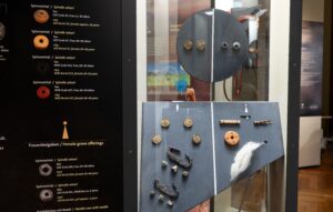 Vitrinen- oder Museumsdisplay mit mehreren archÃ¤ologischen FundstÃ¼cken, darunter runde Spinnwirtel, kleine Metallobjekte, Perlen und eine weiÃe Faserprobe; links eine zweisprachige Infotafel mit Beschriftungen zu den Funden. (Text KI-unterstÃ¼tzt generiert)